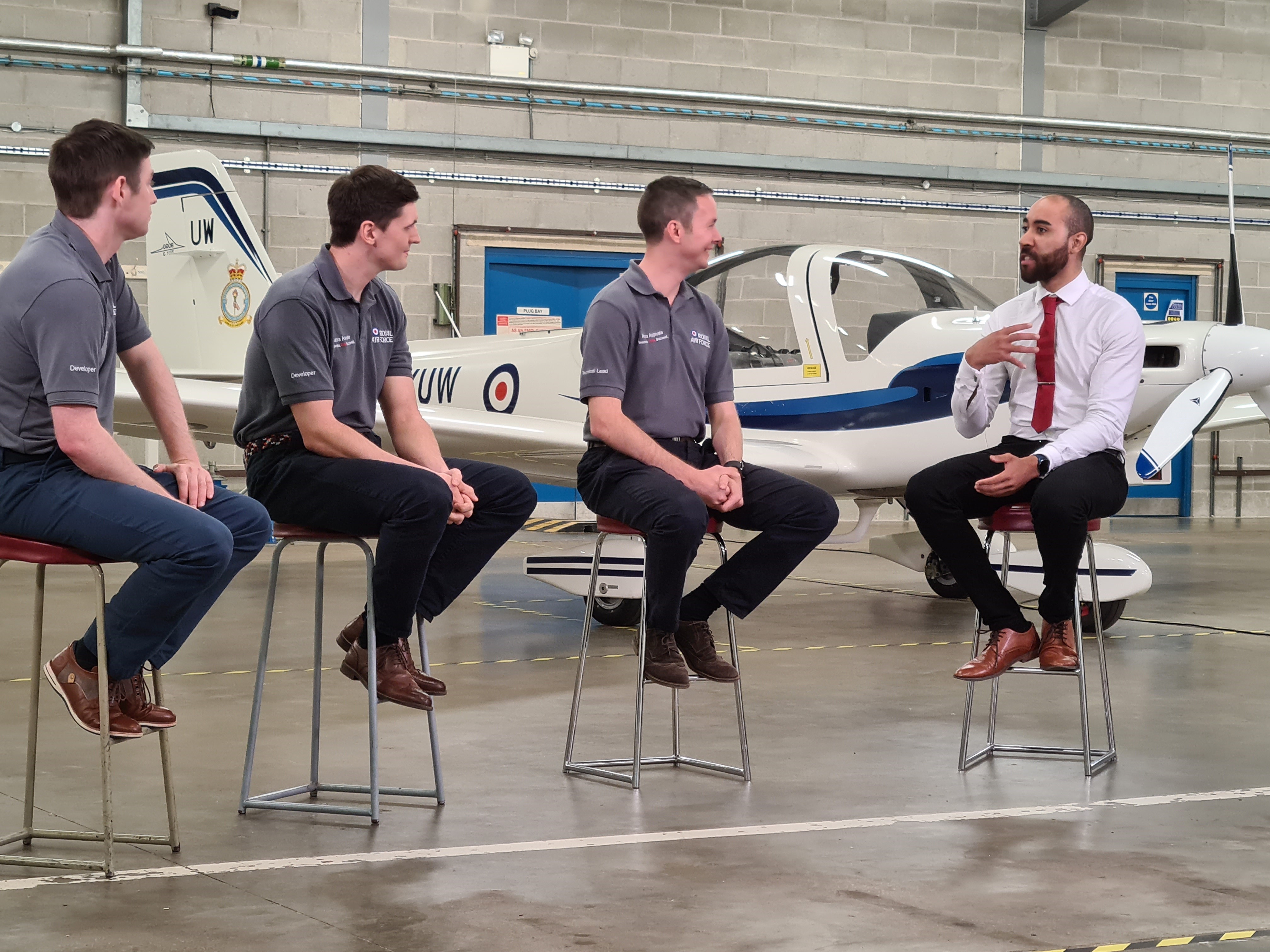 Personnel sit on stools in an aircraft hangar, with interviewer.